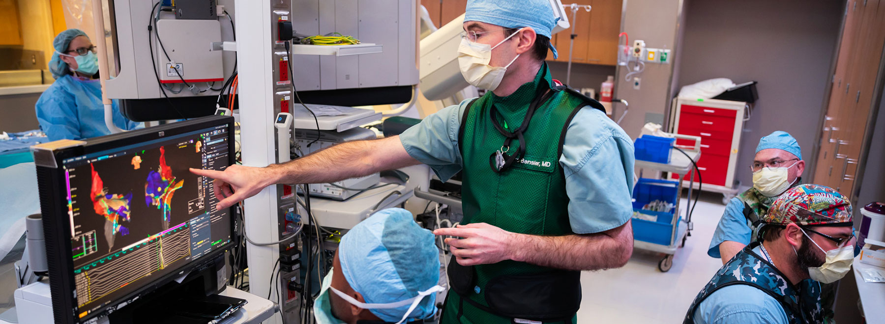 Doctor points at heart imaging screen while medical team observes in procedure room. Doctor points at heart imaging screen while medical team observes in procedure room.